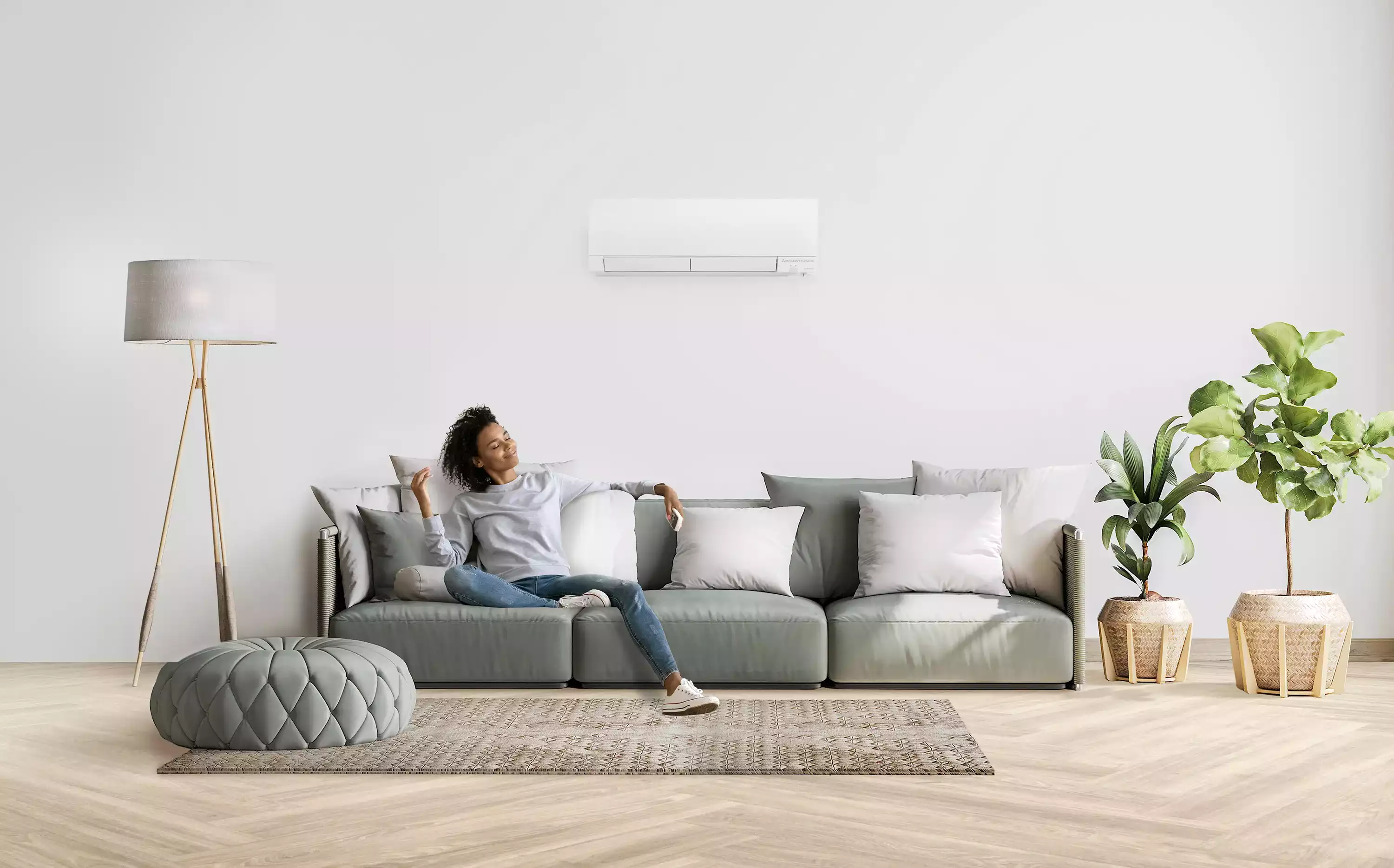 Woman relaxing on a sofa, holding a remote, beneath a wall-mounted air conditioner in a bright living room.